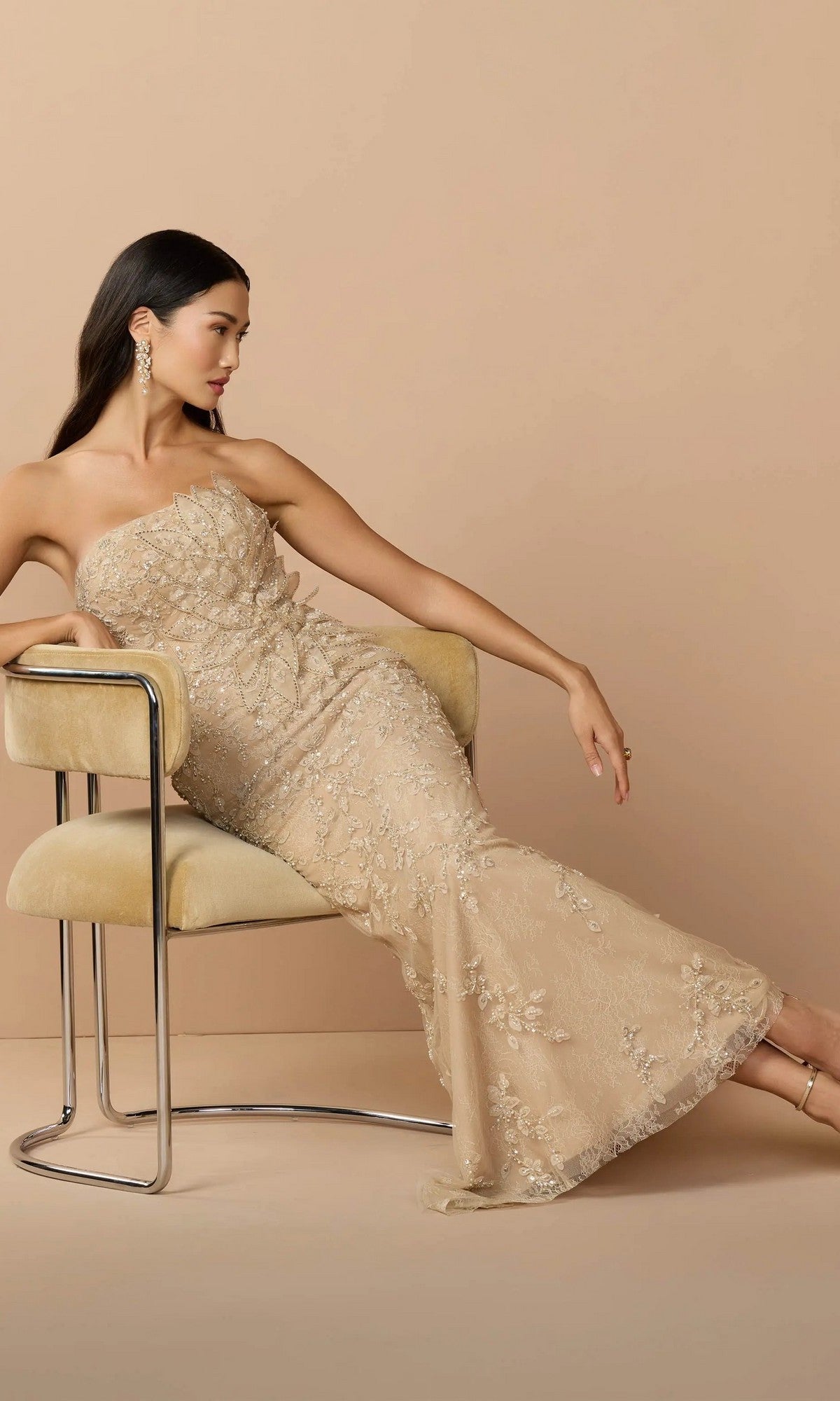 A woman wearing the Mac Duggal 21200 long formal dress in gold sits sideways on a beige chair against a beige background, looking to her left.